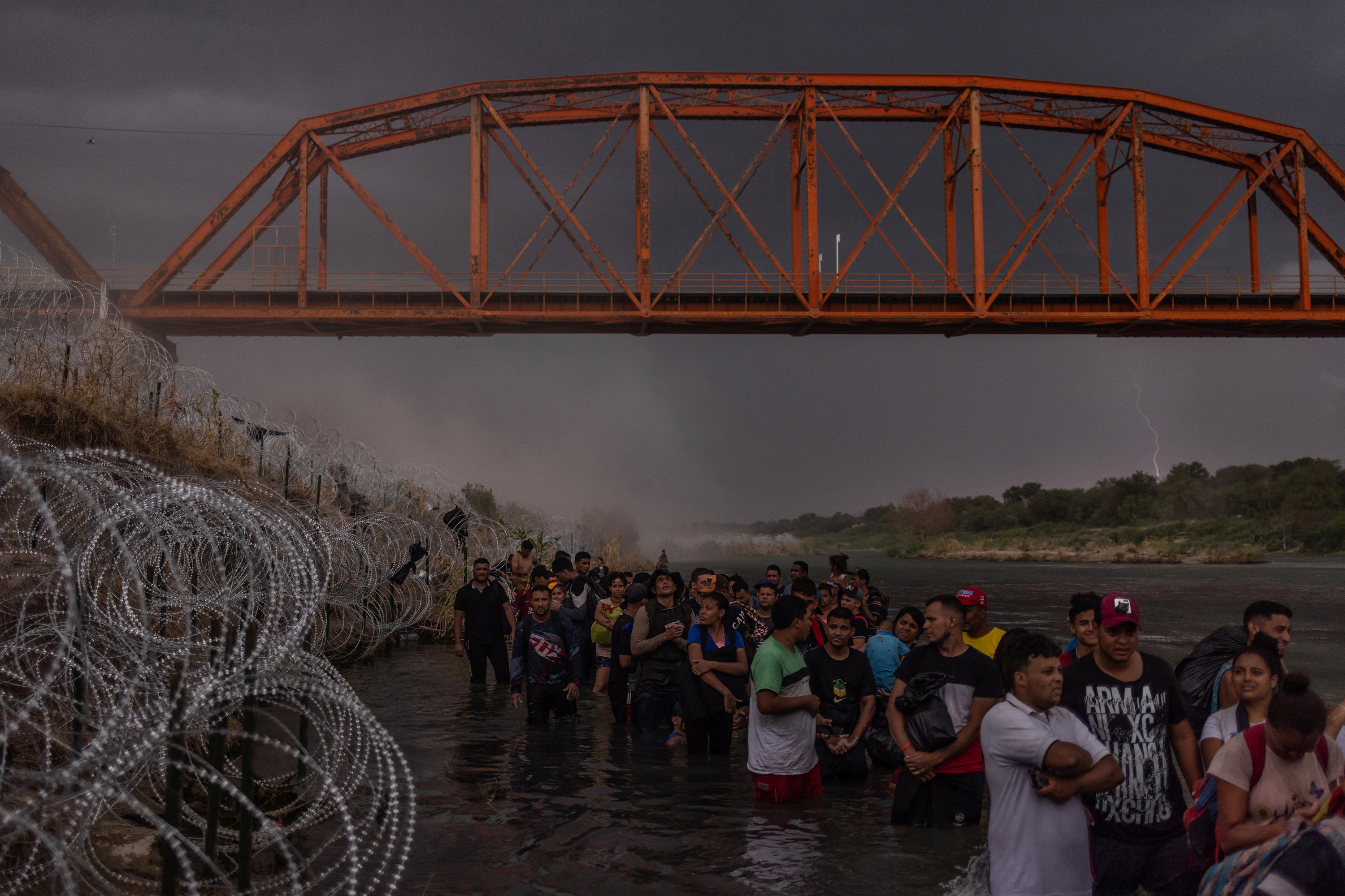 Lighting strikes and dust blows in the background from an incoming thunderstorm as hundreds of migrants line up under the Puente Negro Ferrocarril train bridge in Eagle Pass, waiting to surrender to authorities after wading across the Rio Grande into the U.S. from Mexico on September 15, 2023.